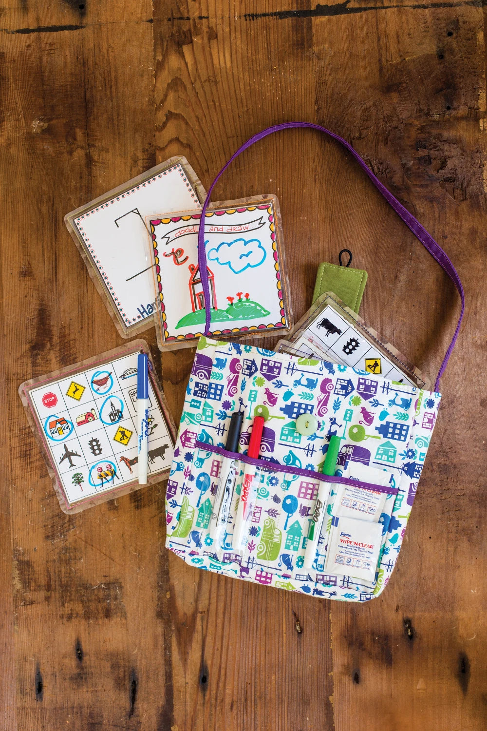A vibrant bag crafted from Quilters Vinyl features a house pattern and holds pens and markers, placed on a wooden table beside four illustrated activity cards with drawings and symbols.