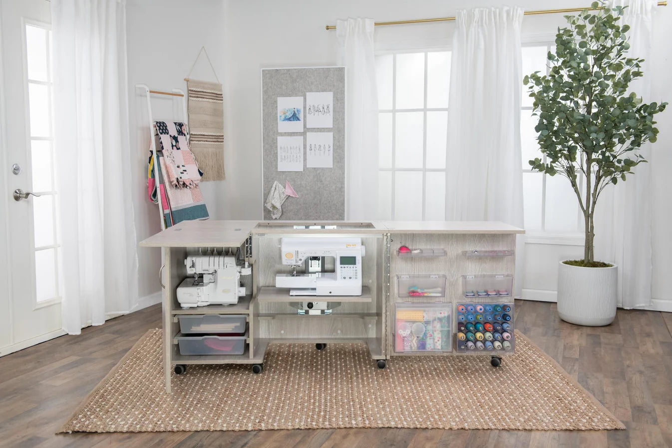 A bright sewing room features the Koala Eclipse Sewing Cabinet in White and Grey Oak with two machines, organized clear bins of supplies, rolls of thread, fabric, a potted tree, wall decor, and white curtains allowing natural light.
