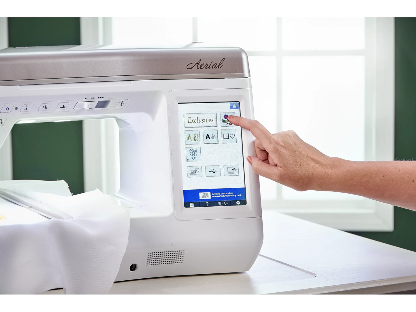A person uses the touchscreen of a Baby Lock Aerial Embroidery and Sewing Machine with 60 days of FREE online classes, guiding fabric under the needle in a bright room with a window.