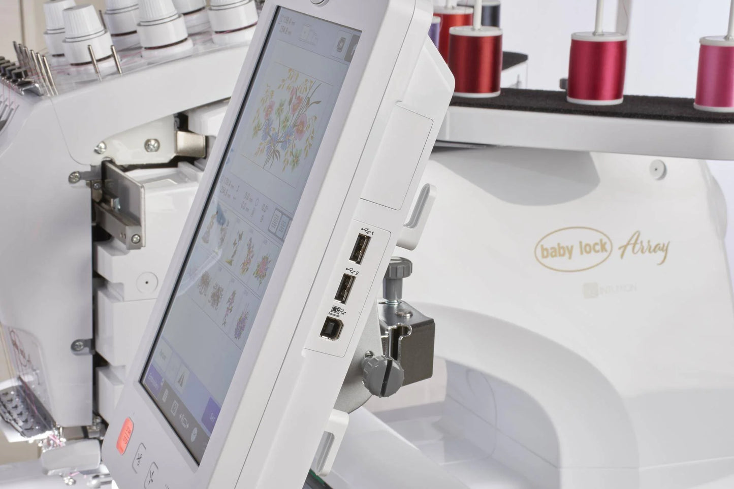 Close-up of the Baby Lock Array 6 Needle Embroidery Machine, featuring its touchscreen display with floral designs, USB ports, and multiple colored thread spools in the background.