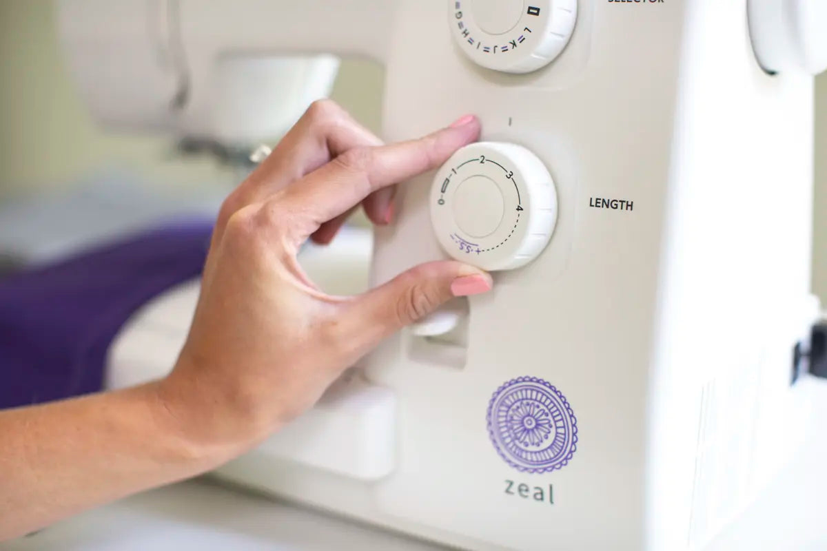 A hand with light pink nail polish adjusts the stitch length dial on a white Baby Lock Zeal Sewing Machine.