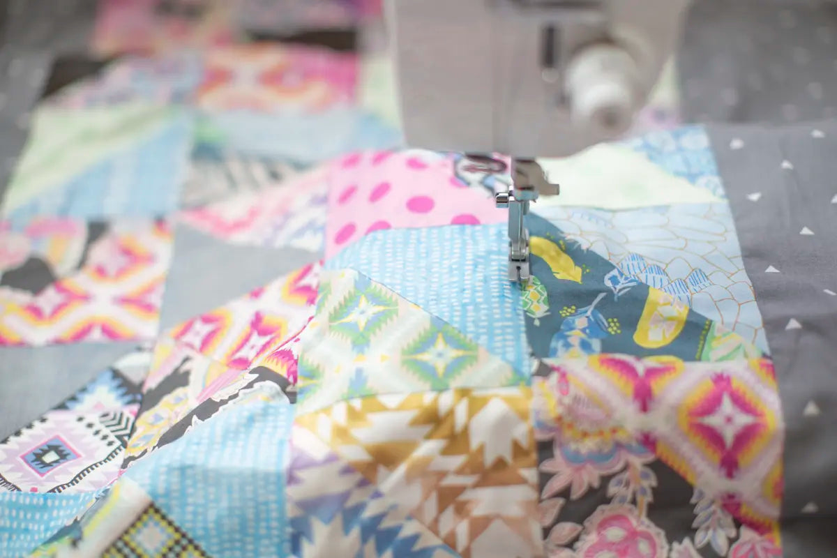 The Baby Lock Accomplish 2 Sewing Machine with FREE online sewing classes stitches vibrant, patterned fabric squares into a colorful quilt, with its needle working through various bright designs during quilting.