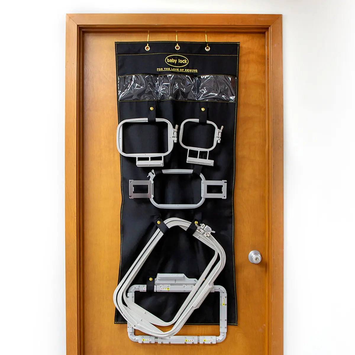 The Baby Lock Over the Door Hoop Hanger is a black organizer with clear and stitched pockets, mounted on a wooden door, providing an efficient way to store rectangular and square embroidery hoops.