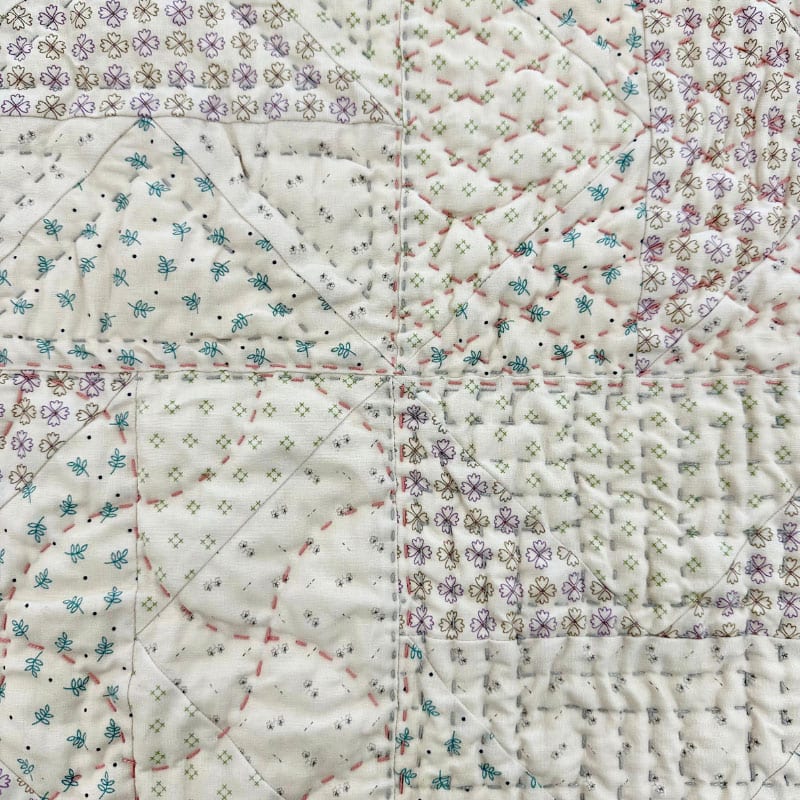 A close-up of a quilt from the Big Stitch Quilting Class, featuring pastel patchwork squares with floral and geometric prints, highlighted by visible cross and diagonal Big Stitch hand quilting in pearl cotton thread.