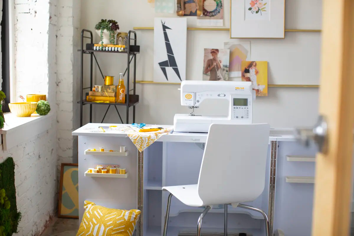 A bright, organized sewing room showcases the Baby Lock Brilliant Sewing Machine on a sleek desk, with yellow and white decor, shelves of supplies, framed art on the wall, and a comfy chair inviting your next creative project.
