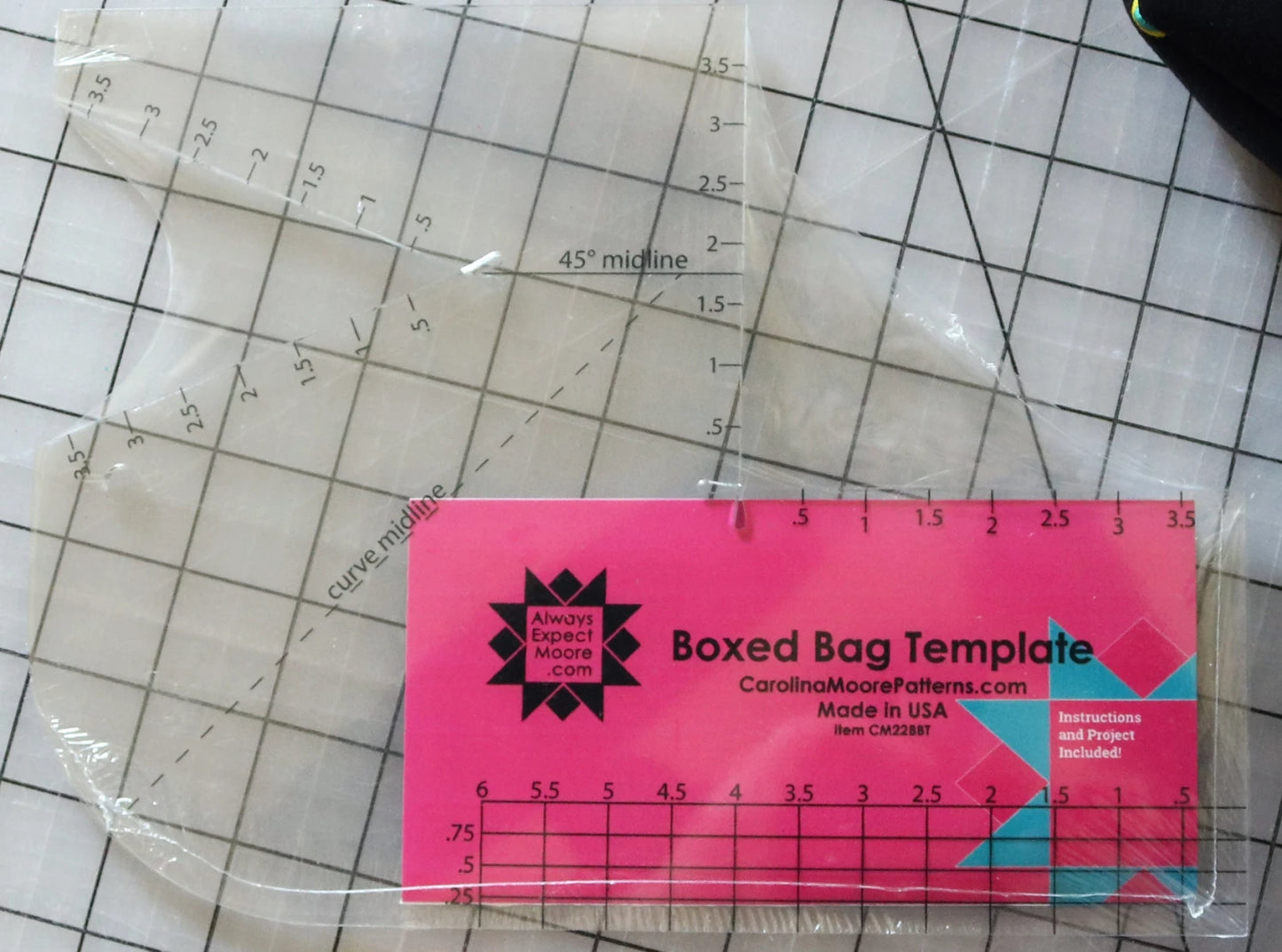 The Checker Distributor Boxed Bag Template, made of transparent acrylic with boxed corners and a pink label, rests on a gridded cutting mat marked for measurements—perfect for sewing projects and use with a rotary cutter.
