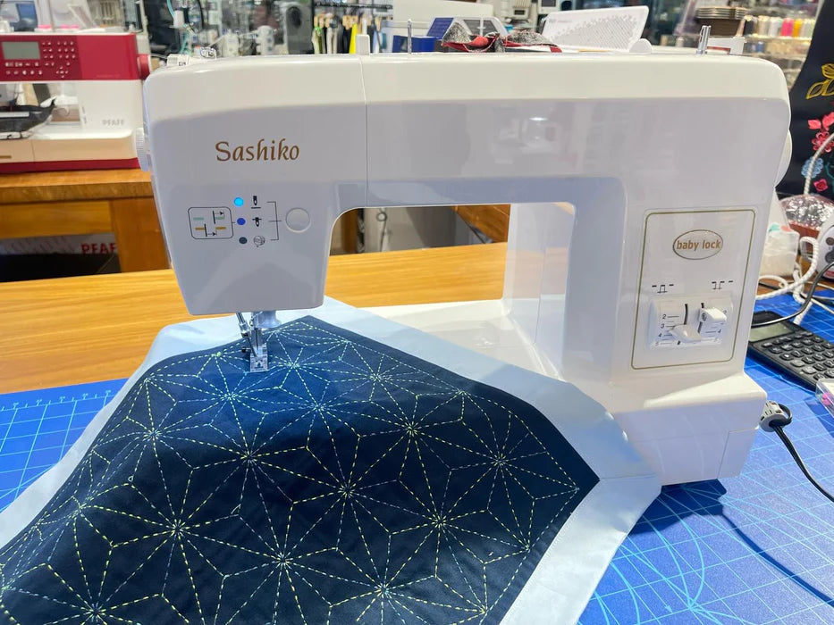 A white Baby Lock Sashiko 2 Specialty Stitch Machine by Baby Lock is stitching a geometric star pattern with yellow thread on dark blue fabric, set on a blue cutting mat in a sewing workspace.