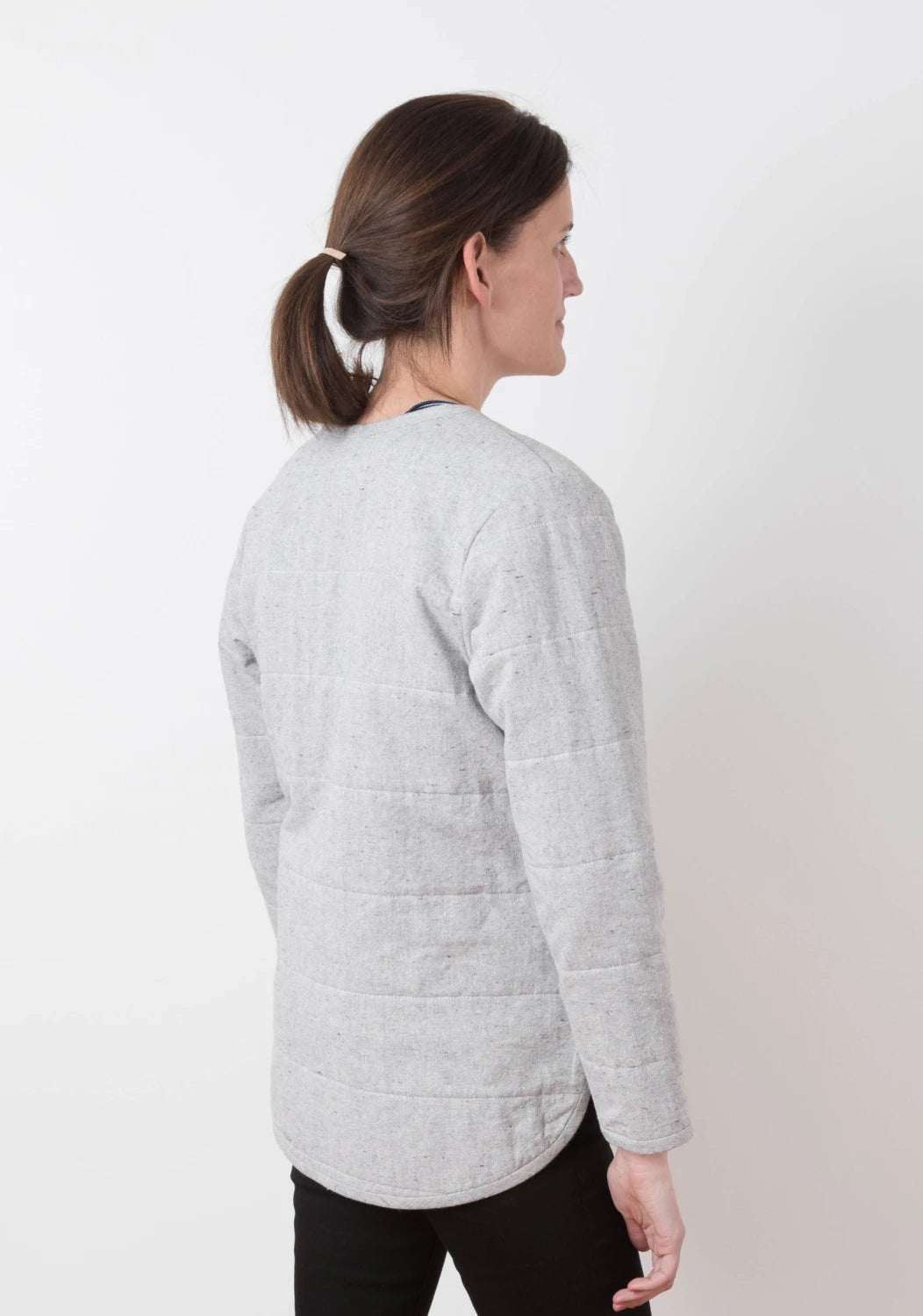 A woman with brown hair in a ponytail models the Checker Distributor Tamarack Jacket Sizes 0-18, a light gray quilted coat perfect for spring and fall layering, paired with black pants against a plain white background.