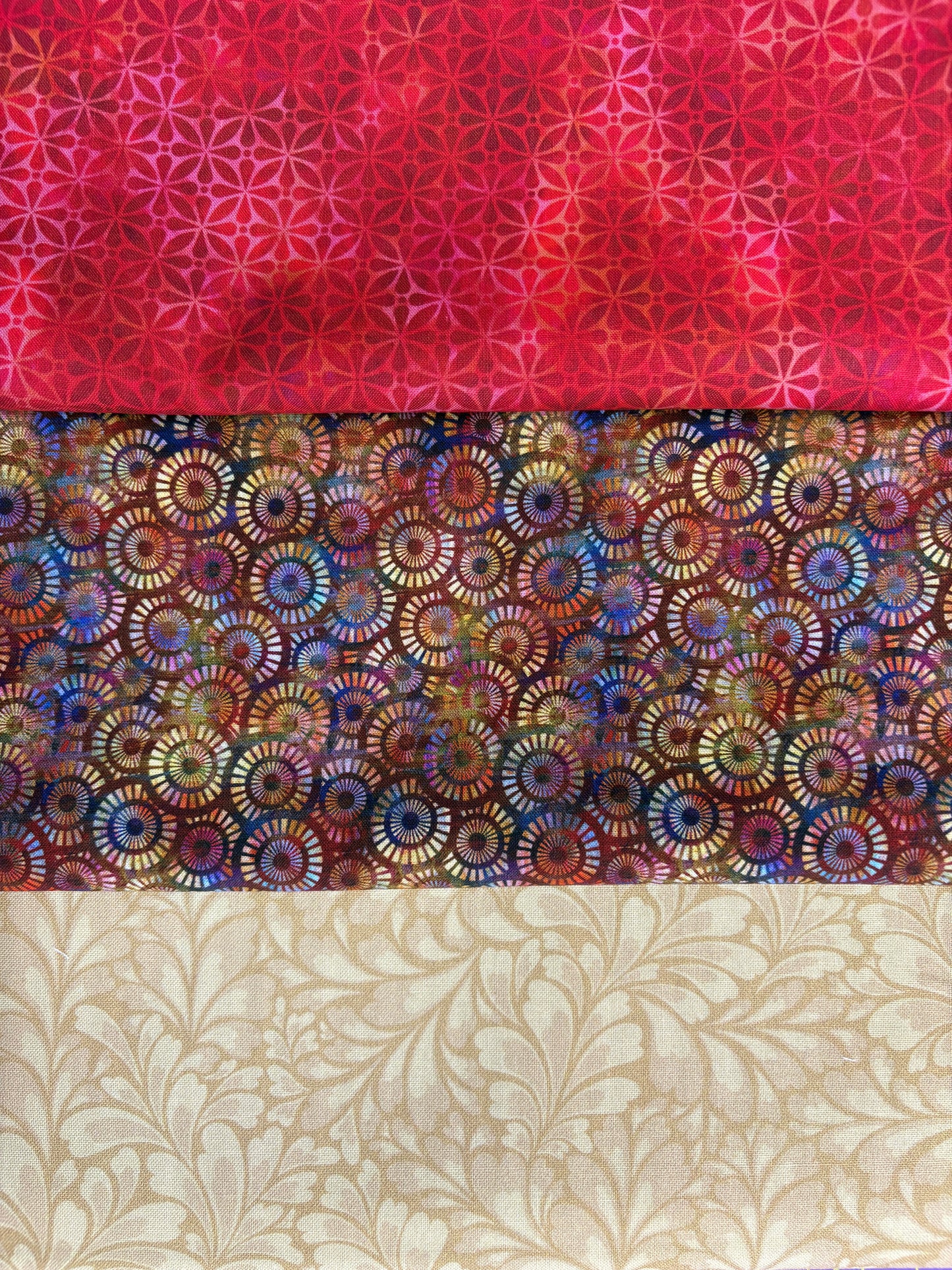 The Gilded Halcyon 3 Yard Quilt Top Bundle by Quilted Joy features a red geometric starburst, a multicolored circular print on dark, and a beige leafy swirl—perfect for pairing with Fabric Cafe 3-yard quilt books. Ideal shop-quality fabric!.