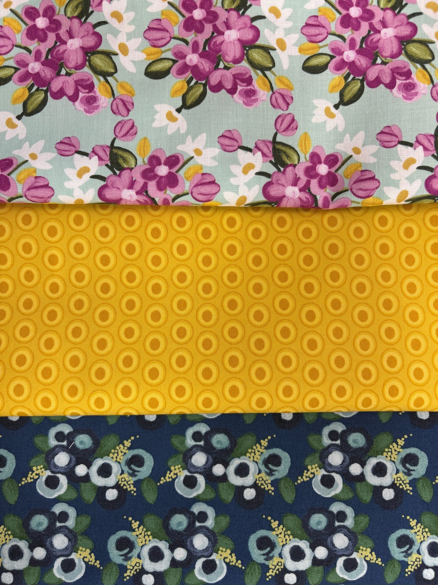 The Quilted Joy Morning Meadow Juleps 3 Yard Quilt Top Bundle includes three patterns: purple and pink flowers with green leaves, yellow with circular motifs, and blue and white flowers on dark blue—perfect for quilting projects.