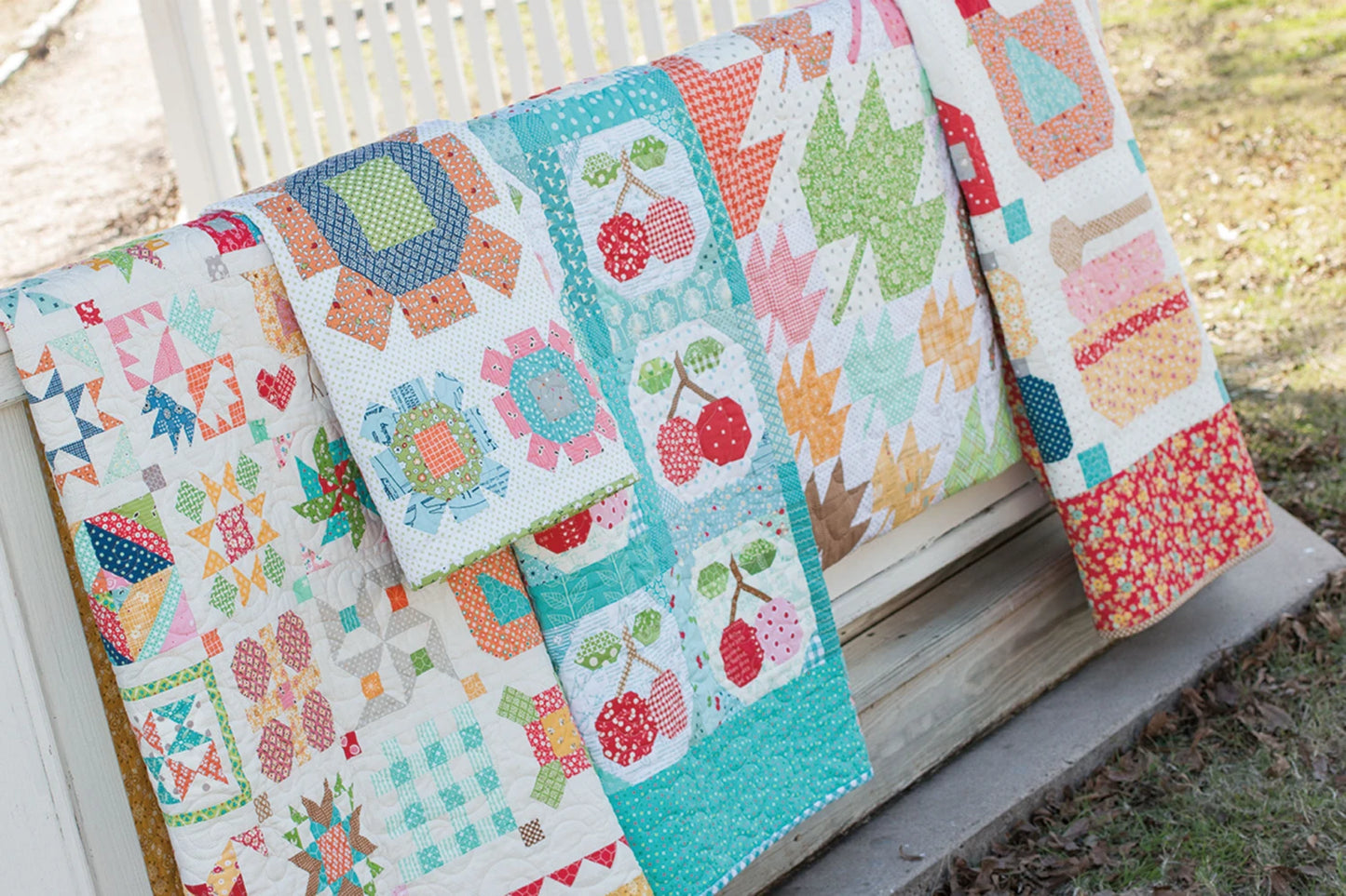 Four colorful quilts, made with Farm Girl Vintage by Checker Distributor, hang on a white fence outdoors. Each features vibrant geometric, floral, and fruit designs. Bright colors stand out near green grass and a sidewalk.