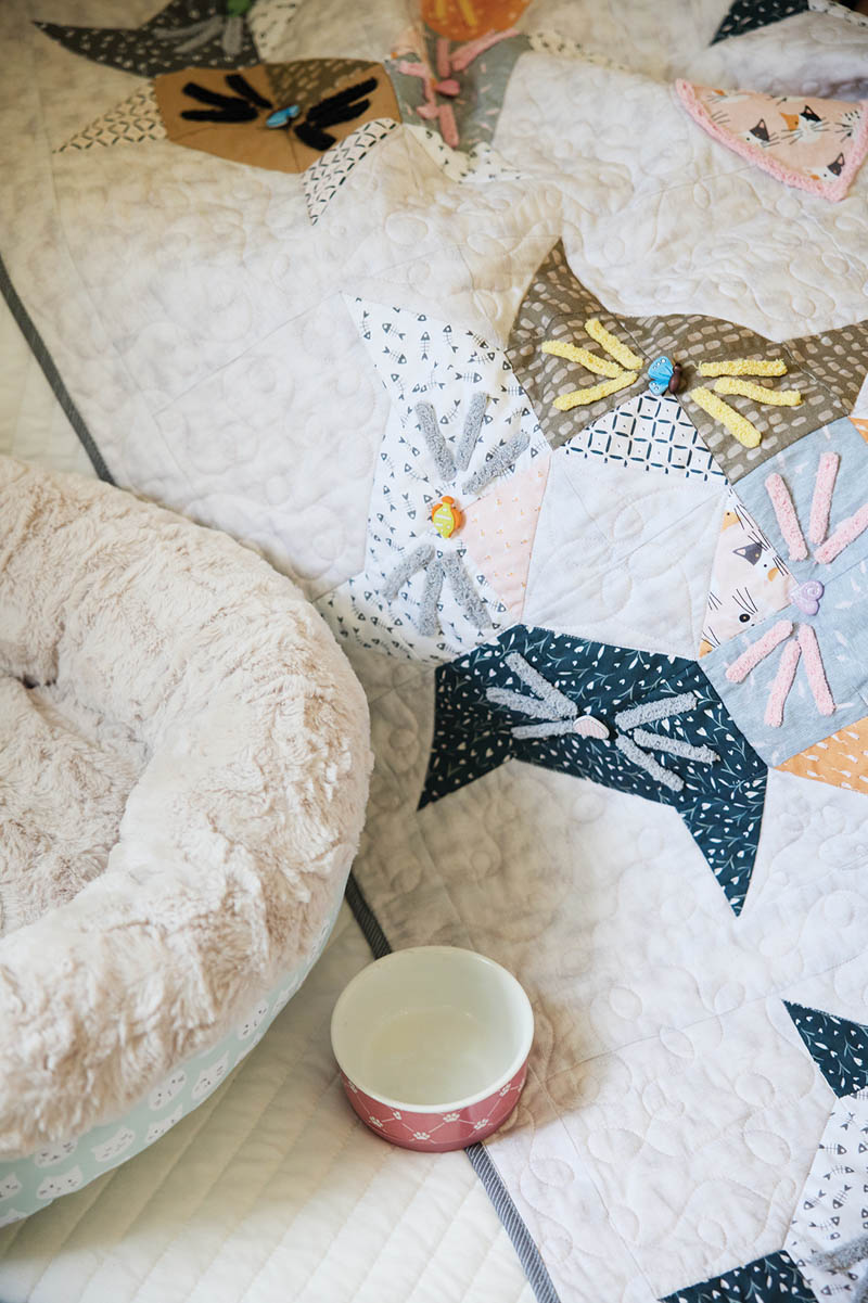 A cozy quilt from the Love of Quilting Fuzzy Love Quilt Kit features colorful patchwork cat faces, draped over a bed next to a plush pet bed and an empty pink bowl on a white quilted surface.