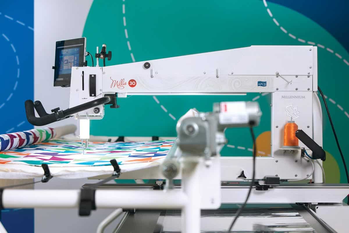 The APQS Demo Millie 30" Long Arm Quilting Machine by APQS stitches a colorful geometric quilt, featuring a digital screen, metal frame, and orange thread spool against a blue and green background.