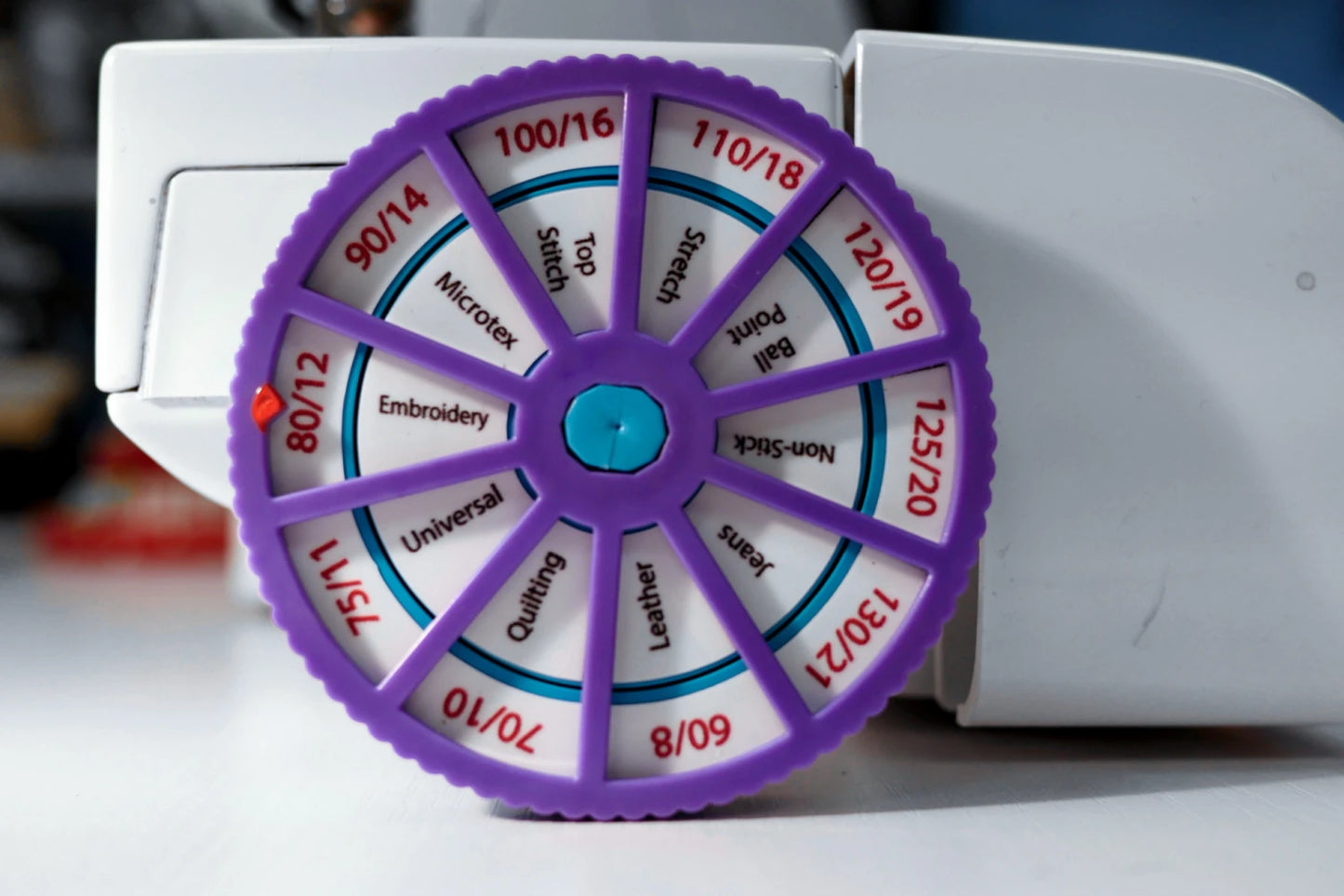 A purple Needle Noter, a circular sewing needle guide, displays embroidery, universal, quilting, and leather sizes and types as it rests against a white sewing machine. This handy accessory helps organize your needles.