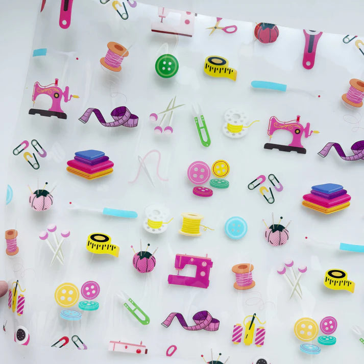 The Clear Vinyl Sew Notion 12"x52" sheet features translucent paper adorned with vibrant sewing-themed prints—thread spools, buttons, tape measures, and more. Perfect inspiration for bag-making projects!.