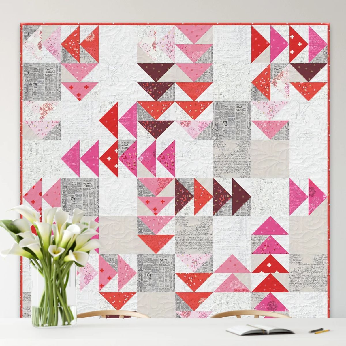 The "My Next Quilt Flying (Spotted) Geese Starts 5/12" quilt by Quilted Joy, featuring red, pink, and purple geometric patterns and precise Flying Geese blocks, hangs above a table with calla lilies and two wooden chairs.