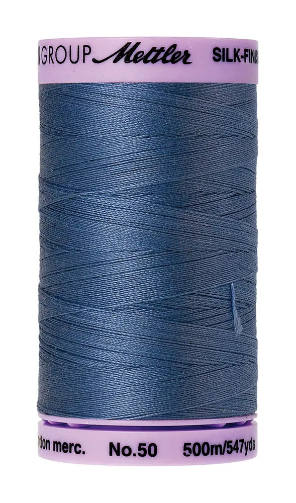 A 500m spool of Mettler Cotton Silk Finish 50wt quilting thread in Smoky Blue, featuring a light purple cap with brand and product details.
