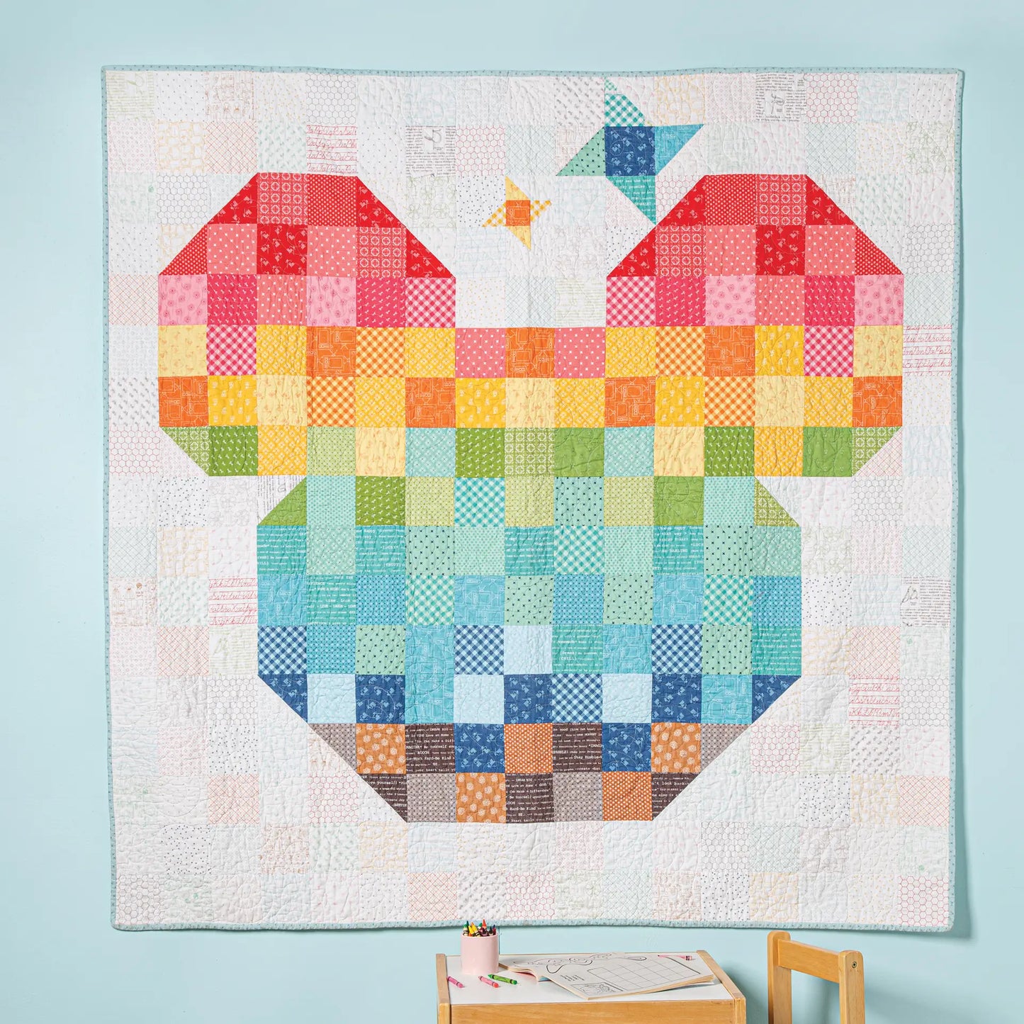 The "Take Me to the Magic Quilt" by Quilted Joy features rainbow geometric shapes forming a bear’s head and star, and hangs on a light blue wall above a small wooden table with crayons and paper.