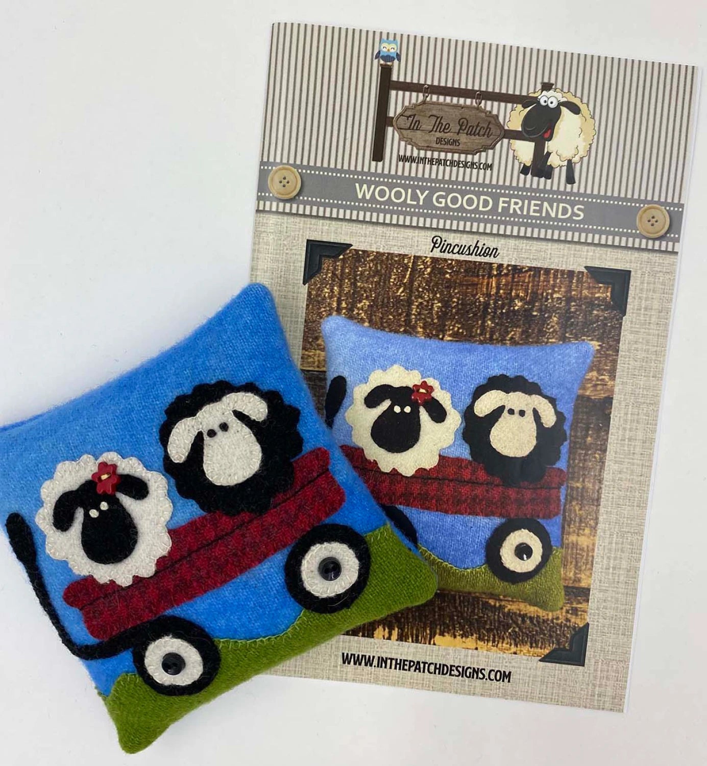 Two handmade pincushions with felt sheep sit in a red wagon on a blue background, displayed on top of the Wooly Good Friends Pincushion Kit from In The Patch Designs.
