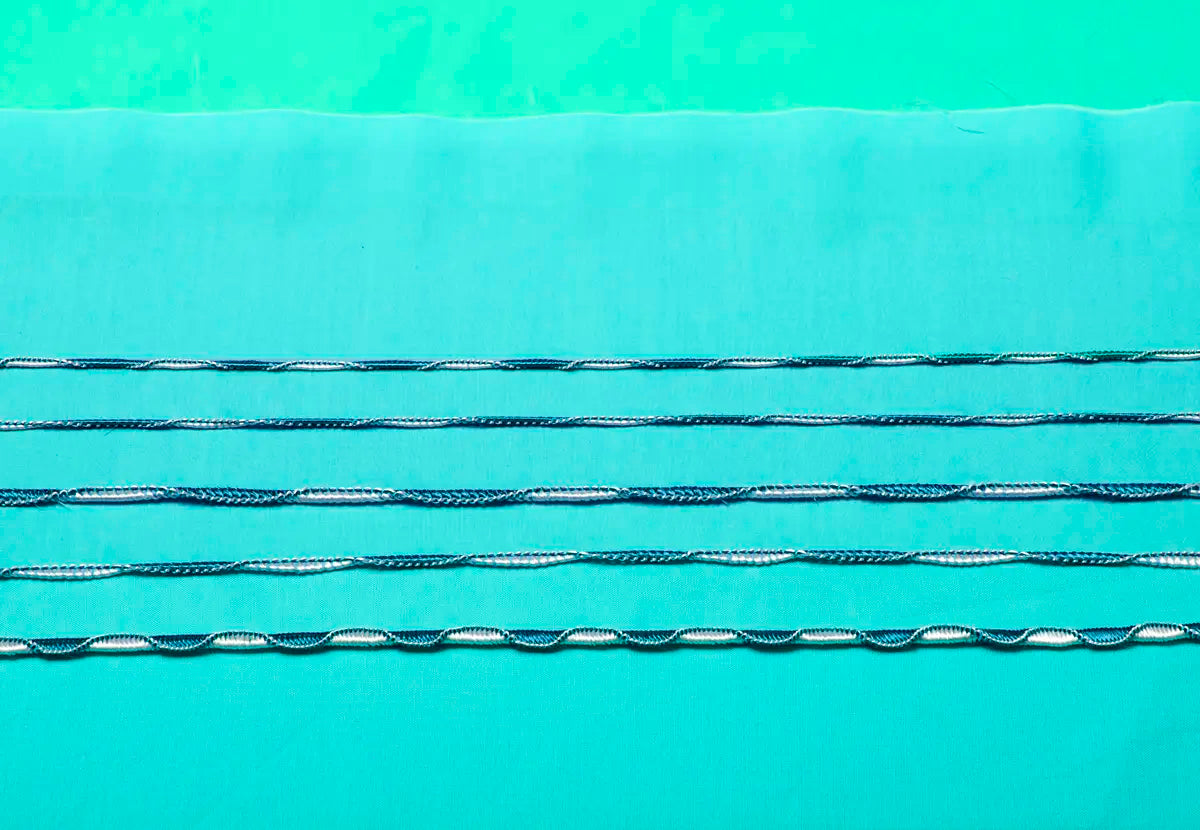 Five parallel rows of surgical sutures in light blue fabric display stitching patterns made with the Baby Lock Triumph Serger Machine, all set on a turquoise background.