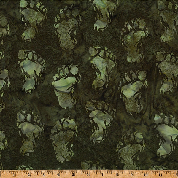 Big Foot Olive Footprints Batik Fabric Yardage by Hoffman Fabrics features dark green and black bear paw prints. A wooden ruler in inches is shown for scale. Ideal quilting cotton for your next project.