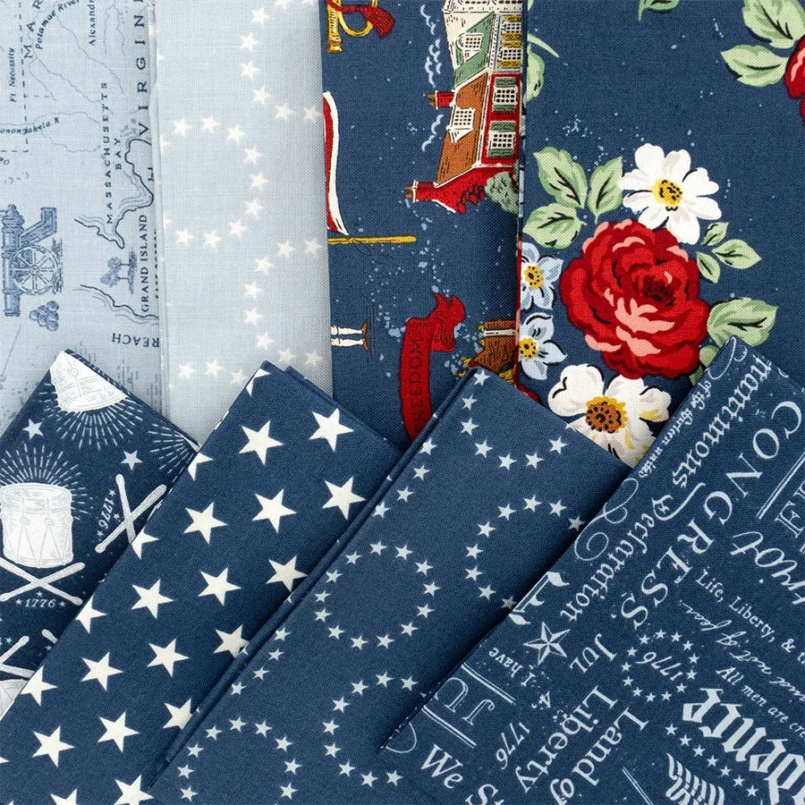 Folded blue fabric pieces from the Riley Blake Independence Fat Quarter Bundle, featuring white stars, cursive text, maps, and red florals, are fanned out on a flat surface—great for quilting projects.