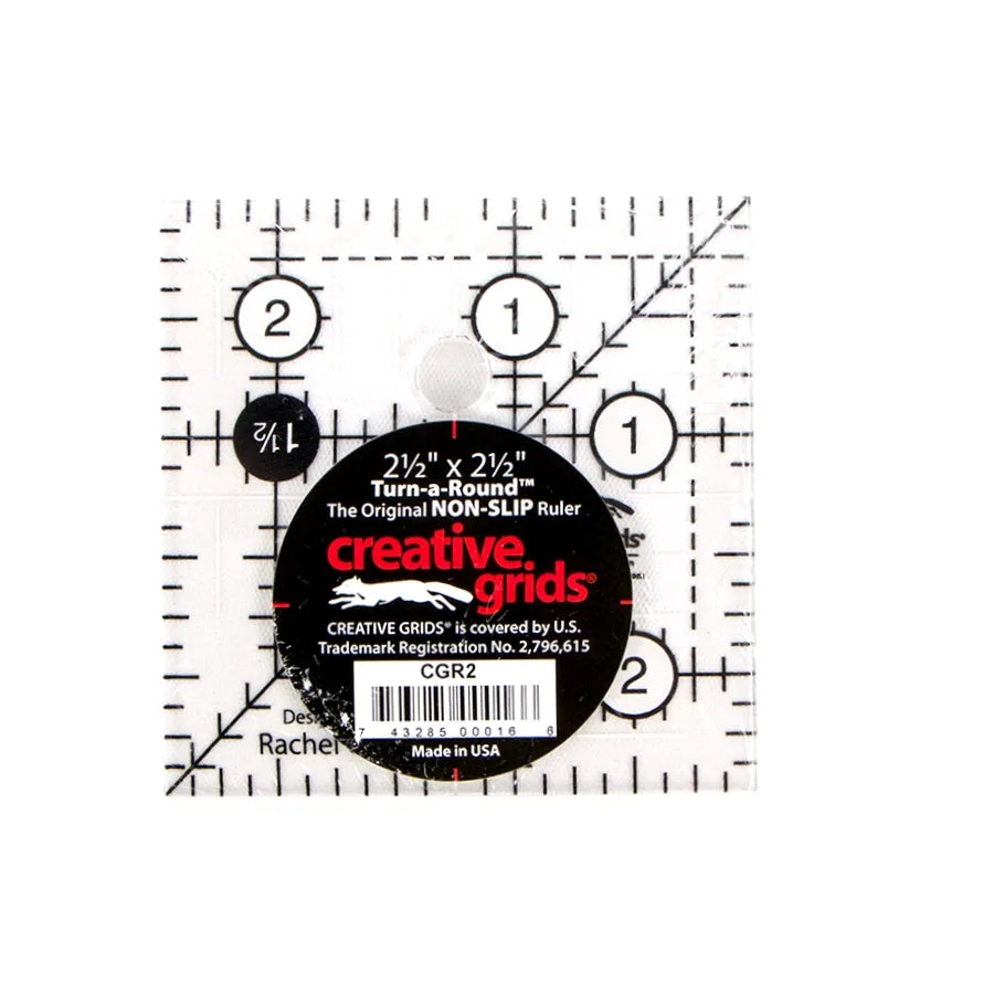 The Checker Distributor Creative Grids Quilt Ruler 2-1/2in Square is a transparent quilting tool with black grid lines, diagonal guides, and circular marks. The center features a black label with bold white and red product details.