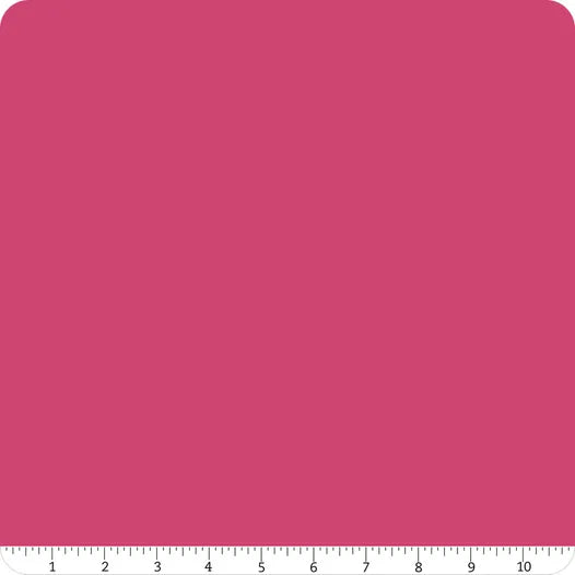 A square swatch of solid fuchsia Confetti Cotton™ Riley Fuchsia Fabric Yardage by Riley Blake, with rounded corners. A white ruler at the bottom edge measures up to 10 inches, showing the size of this quilting cotton sample.