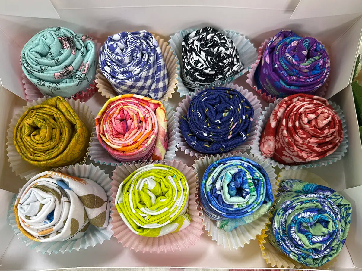 Cupcakes 1 Dozen: twelve quilt shop quality fat quarters with vibrant patterns, each rolled in a paper cupcake liner and arranged in three rows—a delightful sewing gift.