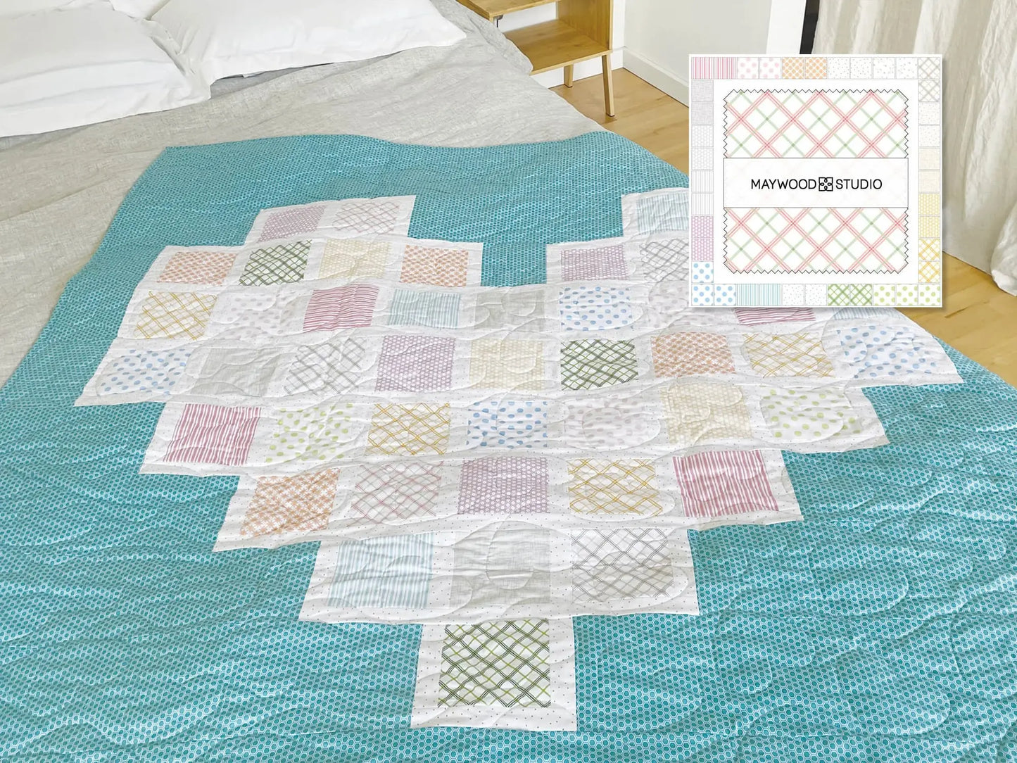 The Kimberbell Full Hearts Teal Quilt Kit creates a heart-shaped quilt displayed over a blue bedspread, with a small “Maywood Studio” box featuring pastel plaid and Lullaby charm squares placed nearby.