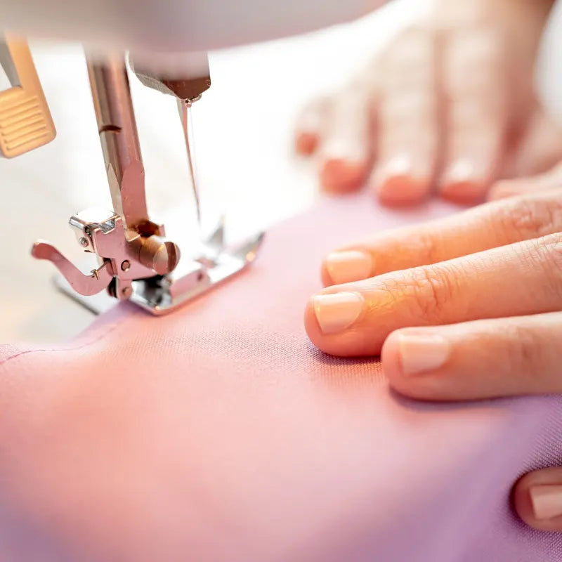 Close-up of hands guiding light pink fabric through a sewing machine, needle and presser foot visible—an ideal scene from the Learn to Use your Sewing Machine Class.