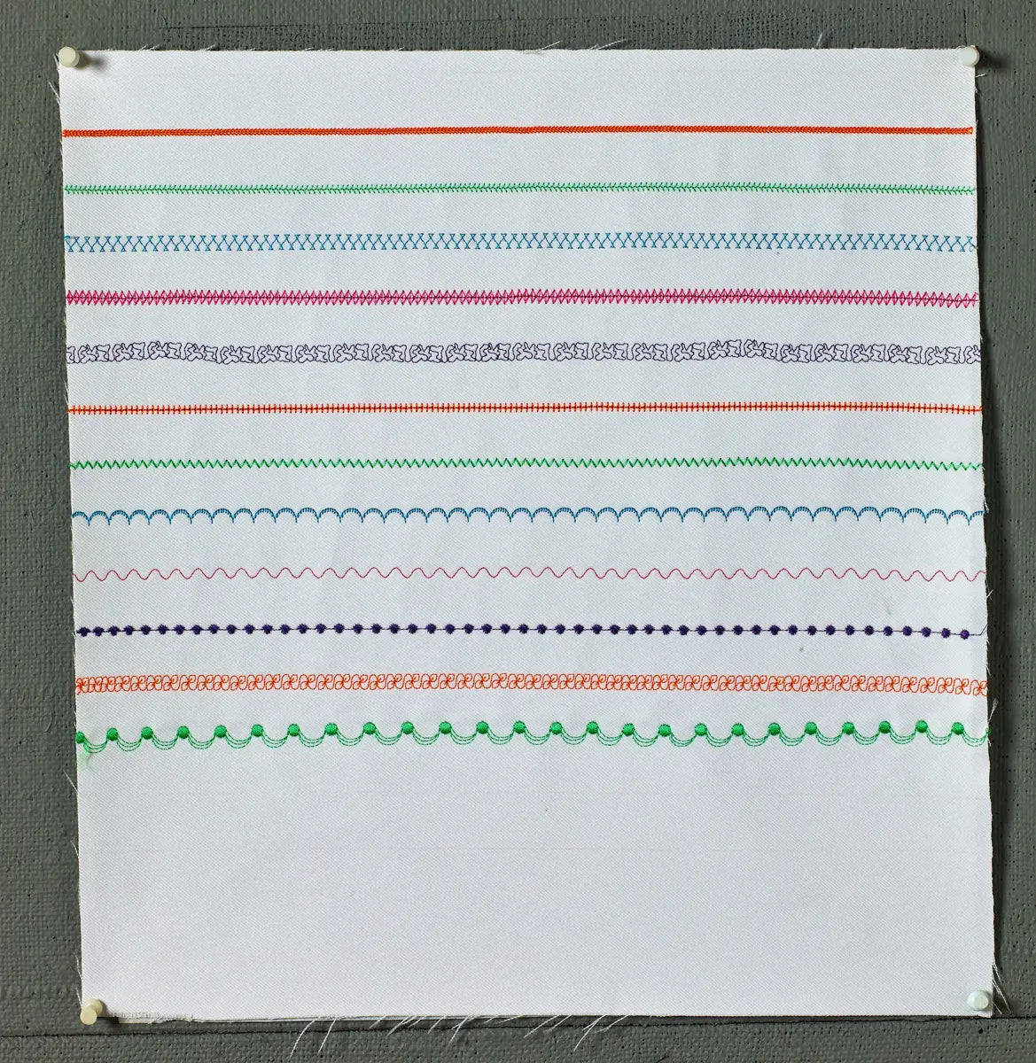 A white fabric square features rows of colorful, decorative built-in stitches—zigzags, scallops, loops—demonstrating the versatility of the Baby Lock Lyric Sewing and Quilting Machine, pinned on a textured gray background.