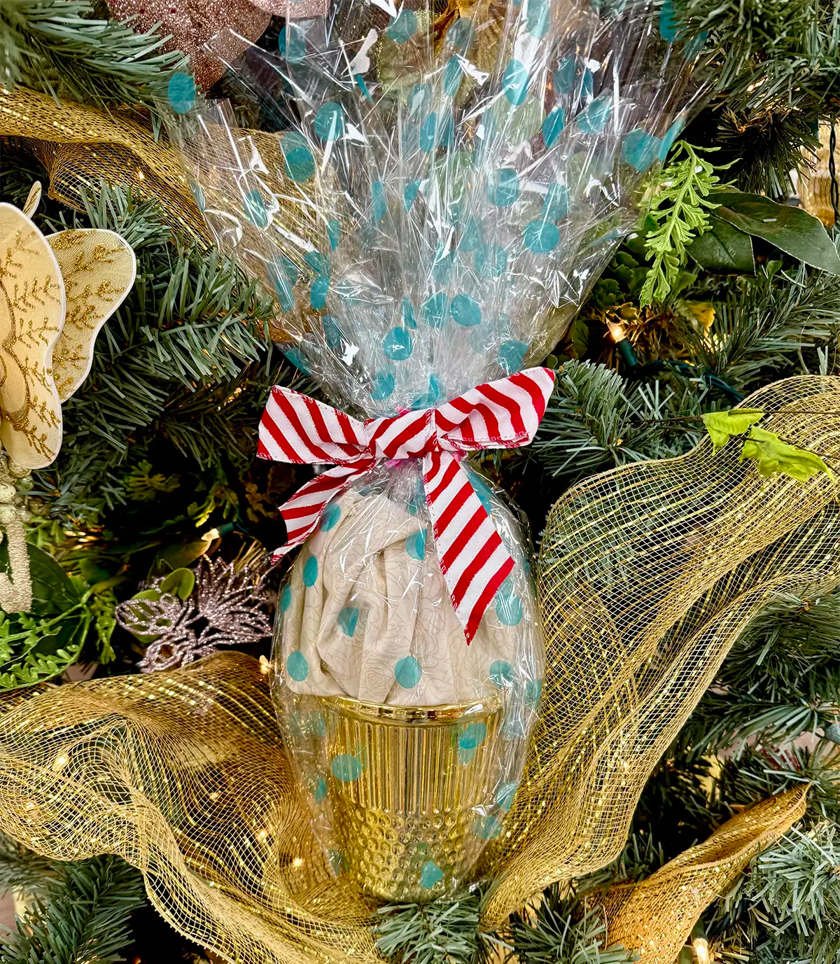 A gold cup wrapped in clear cellophane with teal dots and a red-and-white ribbon rests among pine branches—just like Quilted Joy's Limited Edition Quilt Fairy Surprise Thimble, the perfect quilting gift box for the holidays.