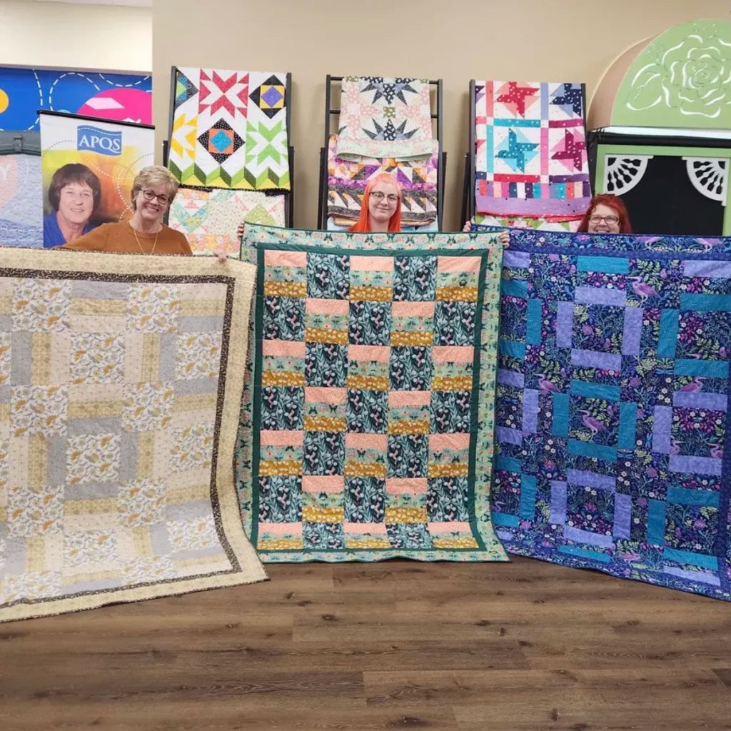 Three smiling women each hold up colorful handmade quilts in front of them, with more displayed on racks behind. The setting is a quilt shop—ideal for those interested in the "My First Quilt Class - Day.