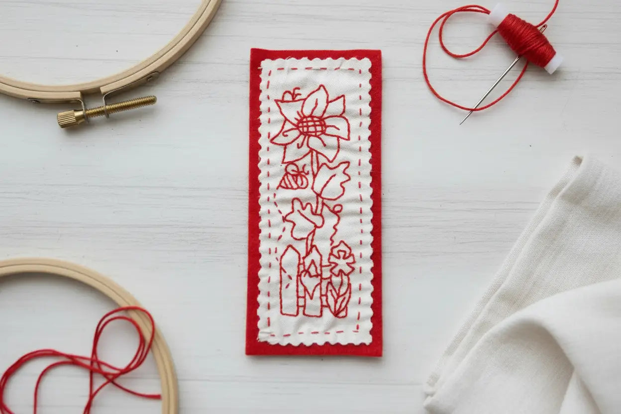 Quilted Joy’s Redwork Hand Embroidery Class showcases handmade bookmarks with red floral embroidery on white fabric, bordered by red felt—surrounded by hoops, red thread, and a needle—a lovely display of basic stitches.