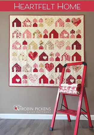 A colorful quilt with red and pink heart and house designs hangs above a red ladder holding quilts. Text reads "Heartfelt Home" and “Quilted Joy.” Featuring the Heartfelt Homes Quilt Kit by Quilted Joy.