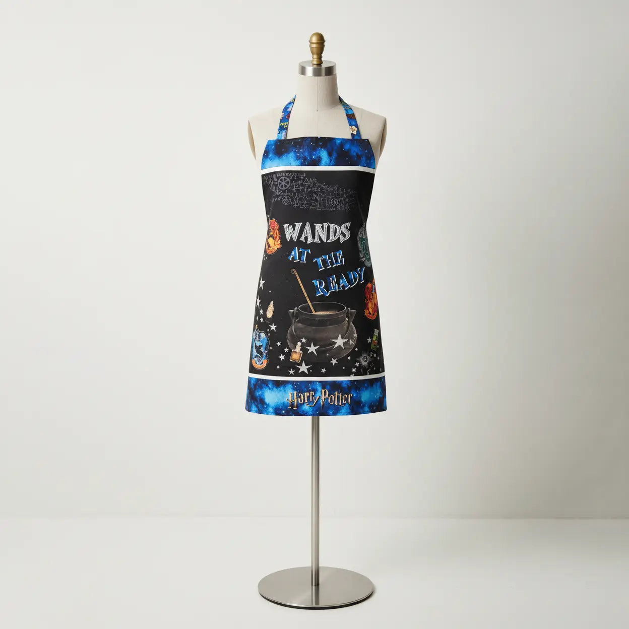 A mannequin displays the Harry Potter Multi Apron Panel by Benartex, featuring "Wands at the Ready," a cauldron, magical symbols, house crests, and blue accents along the edges.