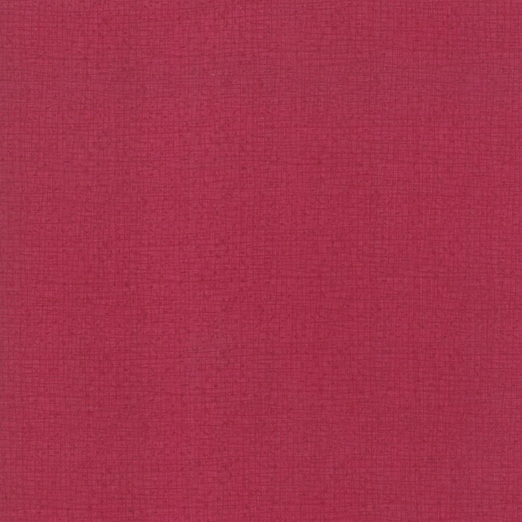 A close-up of United Notions Thatched Cranberry 108" Wide Quilt Backing Fabric displays its textured red surface with a subtle grid pattern and fine woven details.