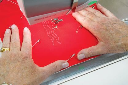 Hands guide red fabric with safety pins under a sewing machine, stitching curves—a perfect moment captured in "Modern Machine Quilting Second Edition" by Checker Distributor for quilting enthusiasts.