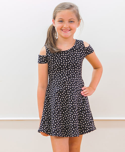 A young girl with brown hair in a side ponytail smiles while wearing a black dress with white polka dots and cold shoulder sleeves. She stands against a plain light-colored background with one hand on her hip.