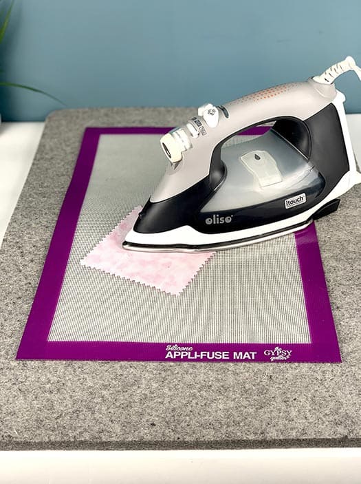 image of an iron pressing on a pink square of fabric, laying on a silicone appli-fuse mat on a grey wool pressing mat