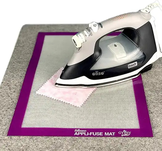 A steam iron rests on a small piece of fabric placed on a mesh applique mat with a purple border labeled Totesew Appli-Fuse Mat on a gray ironing surface.