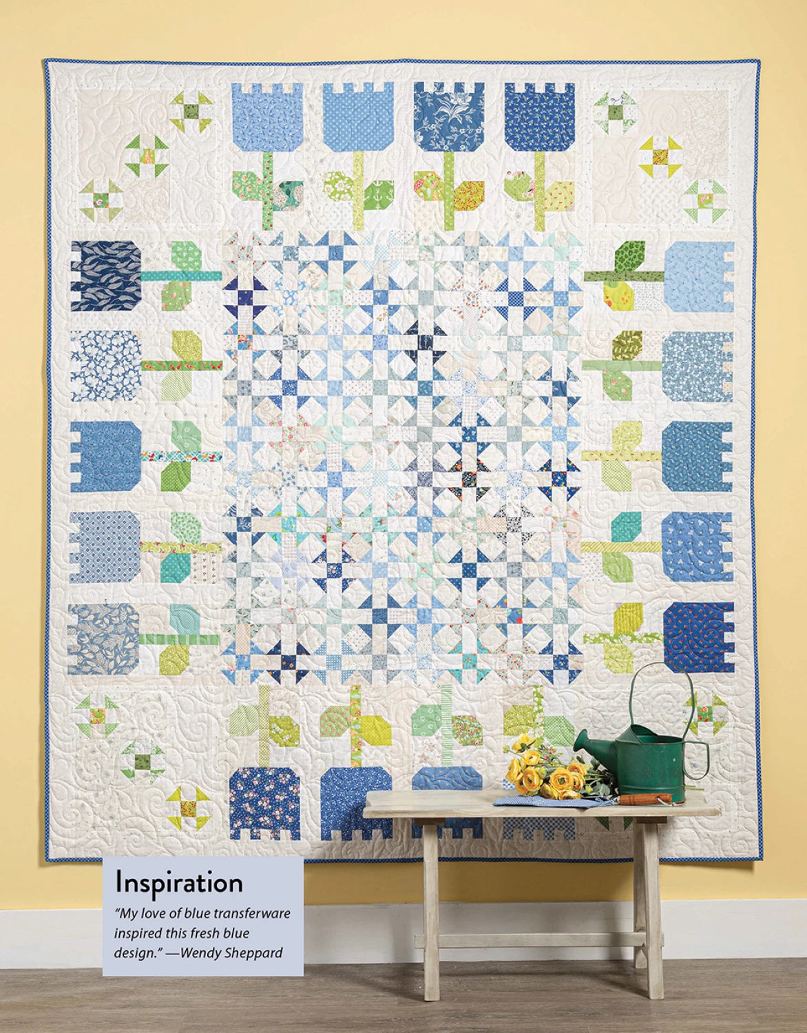 A handmade quilt from Checker Distributor’s "Scrap Quilting With Wendy Sheppard" features blue, green, and white patchwork in floral and geometric patterns. It hangs above a bench with a watering can, yellow flowers, and an “Inspiration” sign.