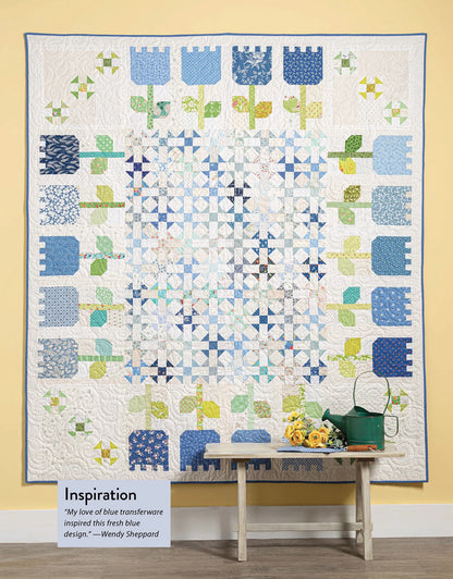 A handmade quilt from Checker Distributor’s "Scrap Quilting With Wendy Sheppard" features blue, green, and white patchwork in floral and geometric patterns. It hangs above a bench with a watering can, yellow flowers, and an “Inspiration” sign.