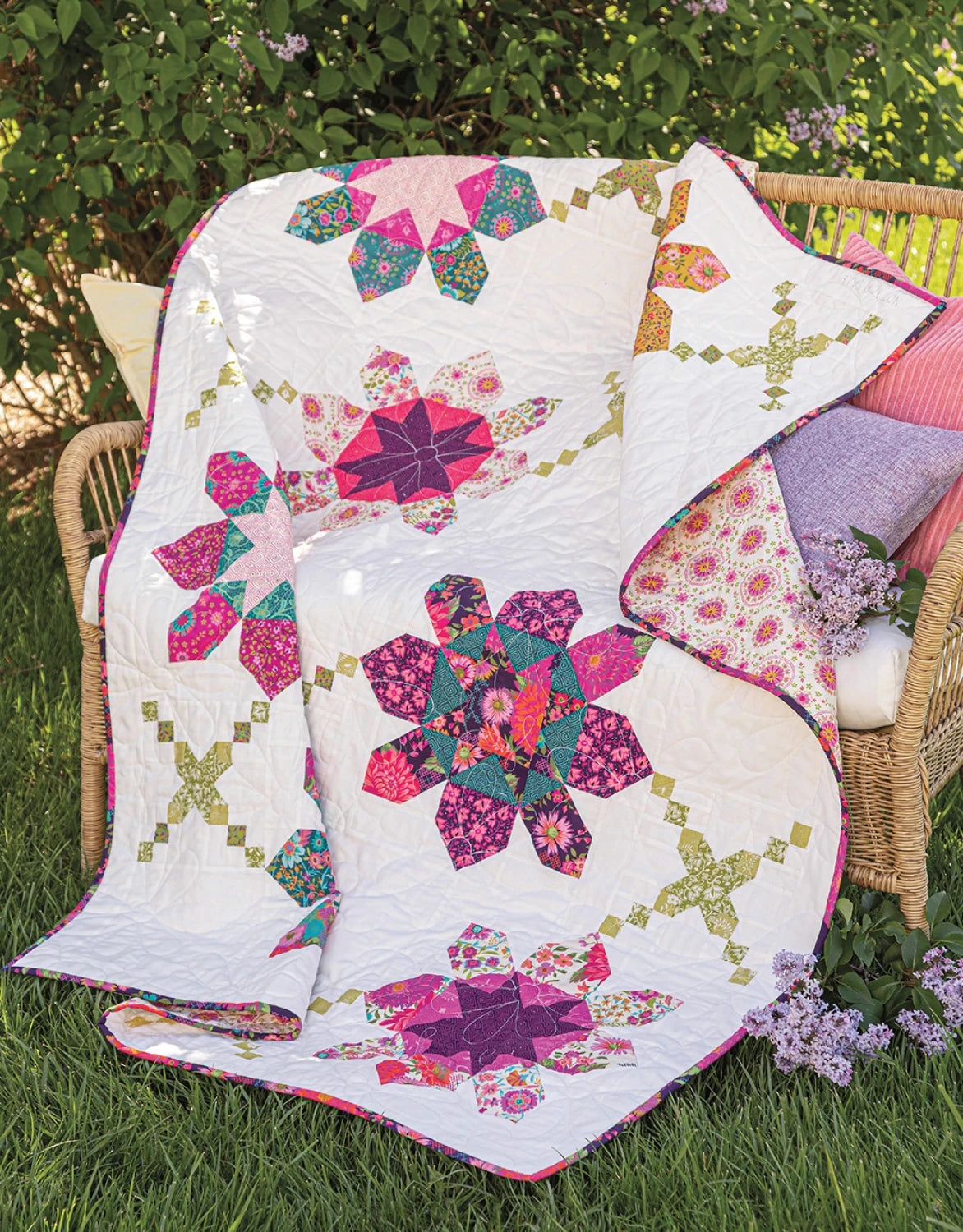 A vibrant quilt made with floral and geometric patterns from "Creative Precut Quilts: Inspiring Designs With Fabric Bundles" by Checker Distributor is draped over a wicker bench with pillows, set on grass near blooming lilac bushes.