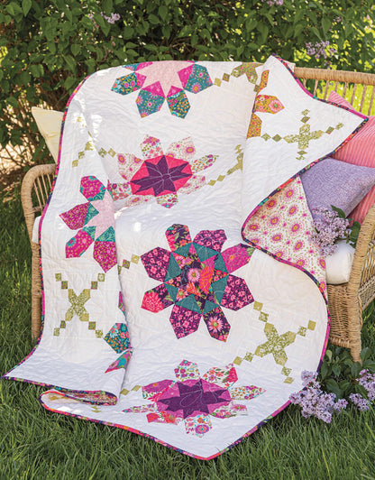 A vibrant quilt made with floral and geometric patterns from "Creative Precut Quilts: Inspiring Designs With Fabric Bundles" by Checker Distributor is draped over a wicker bench with pillows, set on grass near blooming lilac bushes.