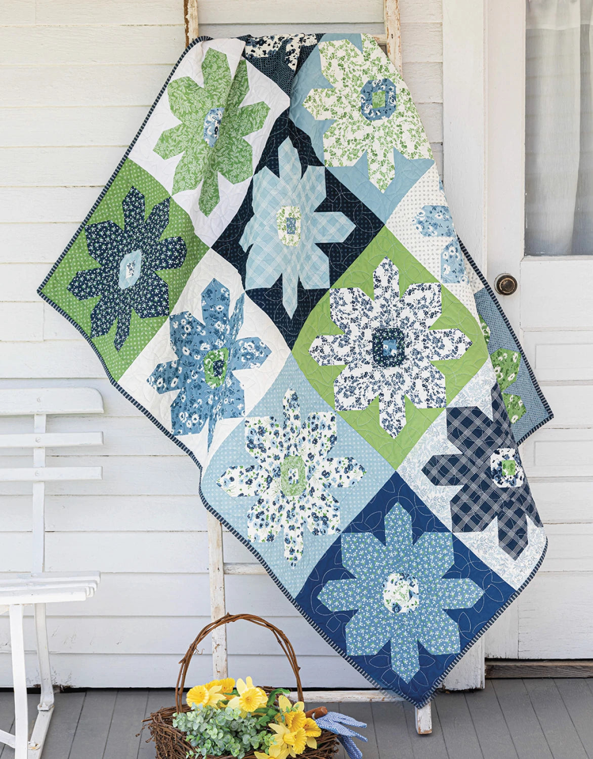 A quilt made from Checker Distributor’s "Creative Precut Quilts: Inspiring Designs With Fabric Bundles" drapes over a white ladder by the door; nearby, a white bench and basket of yellow flowers decorate the porch.