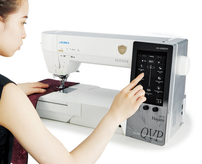 A sewist working at the JUKI Hayate DX-4500QVP, one hand on dark red fabric under the needle and the other selecting on the 7 inch color touchscreen.