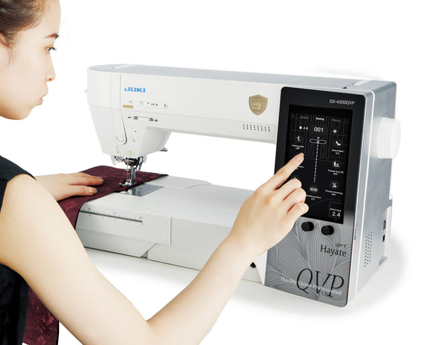 A sewist working at the JUKI Hayate DX-4500QVP, one hand on dark red fabric under the needle and the other selecting on the 7 inch color touchscreen.