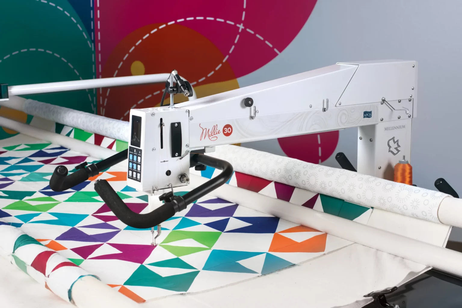 The APQS Demo Millie 30" Long Arm Quilting Machine is quilting a vibrant geometric quilt against an abstract, multicolored background with dotted lines. The white APQS machine features a touchscreen and black handles.